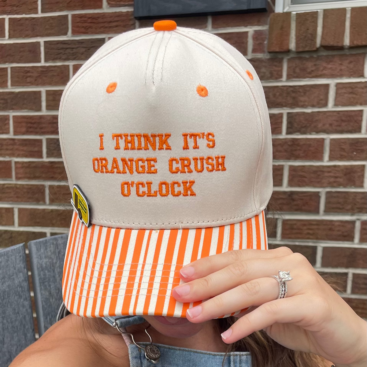 A person wearing a beige snapback cap with orange embroidered text reading 'I THINK IT'S ORANGE CRUSH O'CLOCK' and an orange-and-white striped brim, photographed outdoors in front of a brick wall.