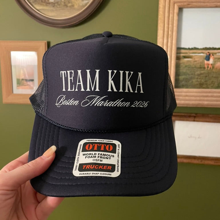 A navy blue trucker hat held by a hand, featuring 'TEAM KIKA Boston Marathon 2026' printed on the front. The hat has a navy mesh back and a white OTTO trucker sticker on the brim, set against a green wall with framed pictures.